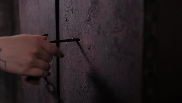 A close-up of a woman's hand with long black nails opens an old rusty metal door with an antique key, a vintage dark room.