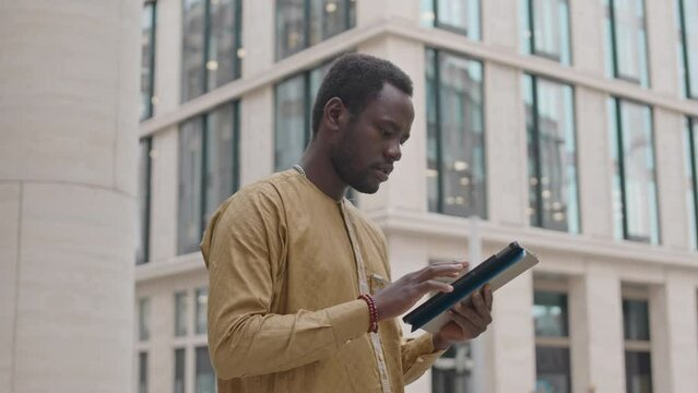 Medium shot of focused young African American man using tablet pc while standing in city downtown with contemporary office building in background