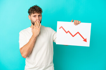 Young handsome caucasian man isolated on blue background holding a sign with a decreasing statistics arrow symbol with surprised expression