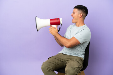 Young man sitting on a chair over isolated background shouting through a megaphone