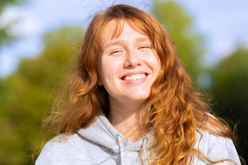 Beautiful young red haired woman in autumn park. Happy ginger girl resting, smiling in nature on background foliage. female portrait broad smile autumn. Lady sunbathing outside on sunset