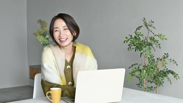 Slow motion video of smiling Asian (Japanese) woman in fall/winter fashion operating laptop in living room Image of home or remote