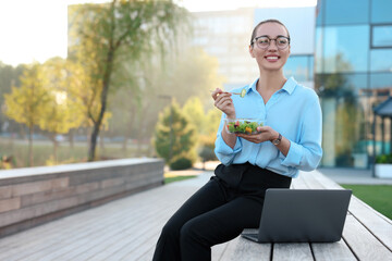 Smiling businesswoman with lunch box sitting near laptop outdoors. Space for text