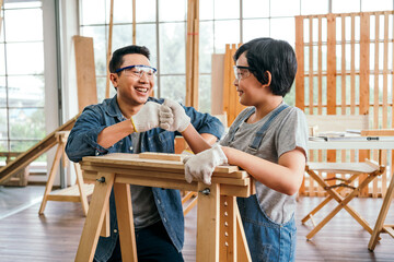 Happy Asian father and son wearing safety glasses and work as a woodworker and carpenter fist bump and smile together after finishing the woodwork project. carpentry working at a home workshop studio.