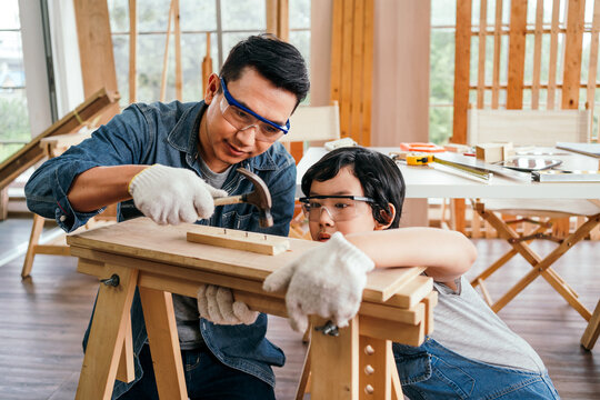 Happy Asian father and son work as a woodworker and carpenter. Father teaching his son to hammer nails on a wooden plank carefully together. carpentry working at a home workshop studio.