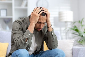 Upset and tired young African American man sitting on couch at home, bowed and holding head in hands. Suffers from depression, panic attacks, thinks about problems