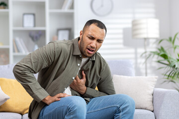 A young African-American man is suffering from a heart attack, sitting on the sofa at home and...