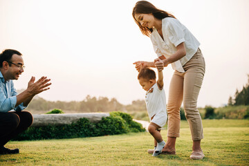 A happy mother and father helping their toddler take her first steps in the green grass of the park. baby learning concept