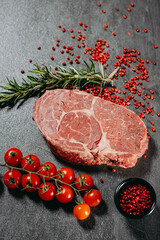 Juicy rib eye steak with red pepper sprinkles in a black saucer, rosemary sprigs and cherry tomatoes next to a kitchen knife on a top view flat lay on gray stone cutting table