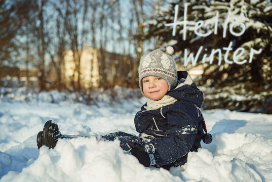 cute boy sitting in snow in park on winter day. Hello winter text on background