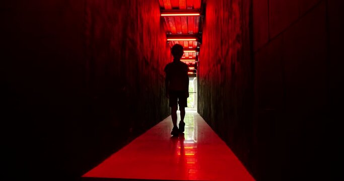 Young boy moves in slow motion through dark corridor illuminated by haunting red light. As he approaches, his silhouette becomes more visible. With impassive face, he creates eerie atmosphere