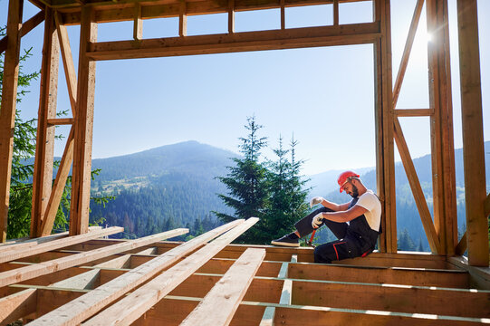 Carpenter constructing wooden frame two-story house near the forest. Bearded man holding hammer, dressed in protective helmet and overalls. Concept of ecological modern construction.