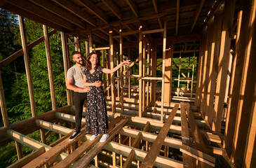 Man and woman appraising their future wooden frame dwelling nestled near the forest. Youthful couple at construction site in early morning. Concept of contemporary ecological construction.