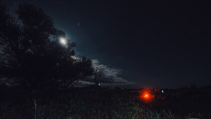 A bright night fire in a camp where a tent is erected and a tree stands. The action takes place at night against the backdrop of the moon and the stars are captured in the photograph.