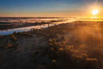Morning sunrise on the big river. The photo was taken from a drone, there is beautiful fog and a blue winding river below.  View of a field next to a river.