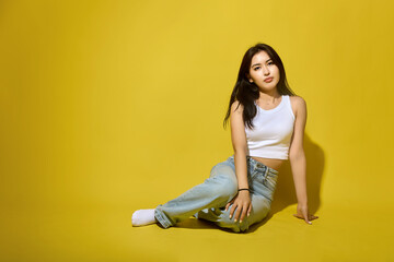 Cute Asian brunette with flowing hair in jeans sitting on a yellow background. Young girl student professional model posing for portfolio