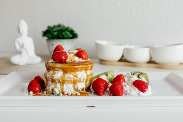 Pouring honey syrup on pancakes. American pancakes with berries and whipped cream on white plate at home. Tasty sweet breakfast food