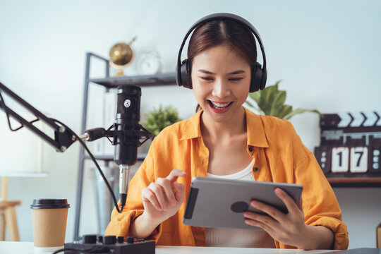 Young Asian woman holding digital tablet and use microphones wear headphones with laptop record podcast interview for radio. Content creator concept.