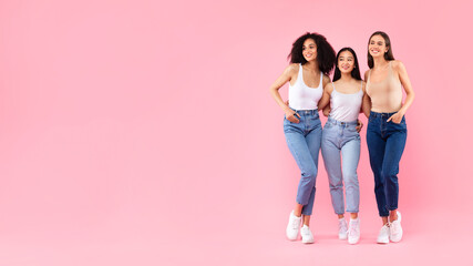 Portrait of three young diverse ladies friends embracing and posing over pink background, panorama...