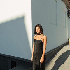 Happy stylish beautiful summer fresh elegant woman with short hairstyle in black fashion spaghetti strap dress walking in the city at sunset. Pretty lady in sunlight outdoors