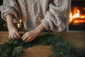 Hands in cozy sweater making Christmas rustic wreath with fir branches on wooden table against cozy fireplace, close up. Winter holiday preparations, atmospheric time