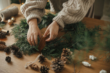 Hands in cozy sweater making Christmas rustic wreath with fir branches, ribbon, pine cones, bells on wooden table, close up. Winter holiday preparations, atmospheric time - Powered by Adobe