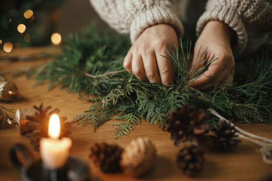 Making Christmas rustic wreath close up. Hands holding cedar branches, making wreath on wooden table with pine cones, candle, bells in atmospheric festive room. Winter holiday preparations