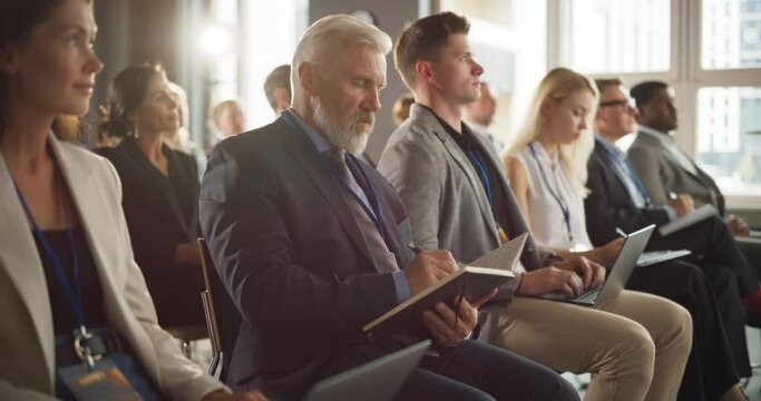 Senior Caucasian Man Sitting in a Crowded Audience at a Business Forum. Corporate Delegate Taking Notes. Successful Male CEO Attending Business Conference, Listening To Presentation.