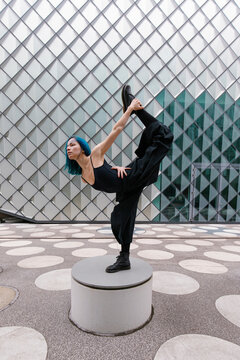 Young flexible dancer dancing on circular concrete seat in front of modern building