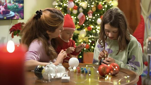 A young attractive mother and her cute daughter and baby are painting Christmas decorations on Christmas Eve, preparing for the celebration and decorating the Christmas tree
