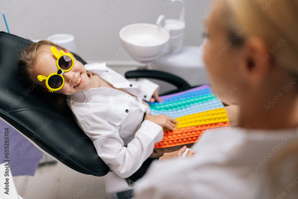 Wall mural side view of cheerful little child girl in funny eyeglasses playing with colorful pop it toy, sittin