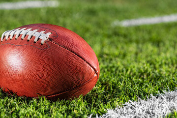 A low angle close-up view of a leather American Football sitting in the grass. It is next to a white yard line with hash marks in the background.