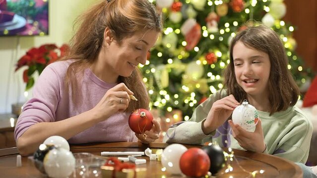 A young attractive mother and her cute daughter are painting Christmas decorations on Christmas Eve, preparing for the celebration and decorating the Christmas tree