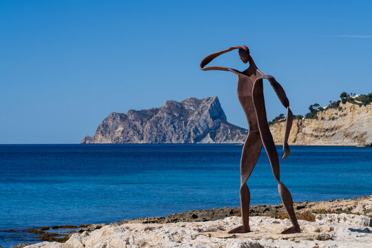A striking iron sculpture of a human figure by Toni Mari on the rocky coast of Moraira, Spain, looking at Calpe's Pe&ntilde;&oacute;n de Ifach across the blue sea.