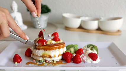 Close-up woman hands cutting pancakes with a fork and knife. American pancakes with berries and whipped cream on white plate at home. Tasty sweet breakfast food
