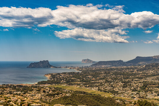 A panoramic view looking south from Cumbre del Sol across Moraira toward the striking Pe&ntilde;on de Ifach rock in Calpe, Spain, beneath a bright blue sky with scattered clouds.