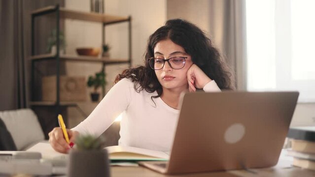 Boring tired young student freelancer with glasses can not study or work making notes at home workplace Stressed curly female exhausted from remote distance work or education and hard program indoors