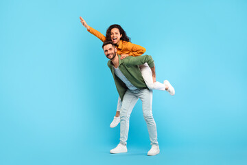 Fototapeta na wymiar Handsome man giving piggyback ride to his wife against blue studio background, full length portrait, free space