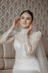 A bride in a wedding dress poses while sitting on a sofa in her room. Portrait of the bride.