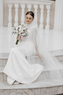 The bride in a long dress sits near white Roman-style columns. Beautiful hair and makeup. An exquisite wedding