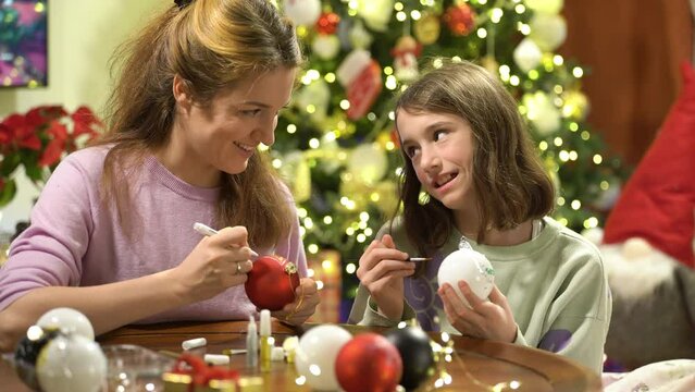A young attractive mother and her cute daughter are painting Christmas decorations on Christmas Eve, preparing for the celebration and decorating the Christmas tree