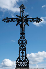 Figure of Jesus Christ on a black painted decorated metal cross, near a gravestone in a cemetery. Clear blue cloudy sky