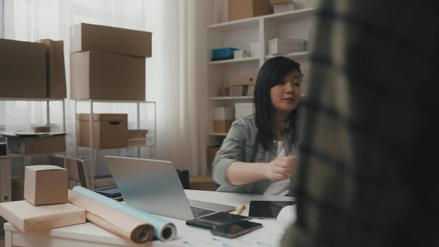 Friendly Asian post office worker giving parcel to customer, working laptop