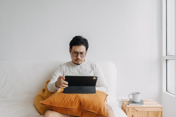 Asian man working using tablet, sitting on sofa at apartment,  thinking something seriously, wearing sweater in cold day.
