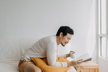 Happy Asian man enjoy reading the book and drinking coffee tea, sitting on sofa at apartment in cold weather.