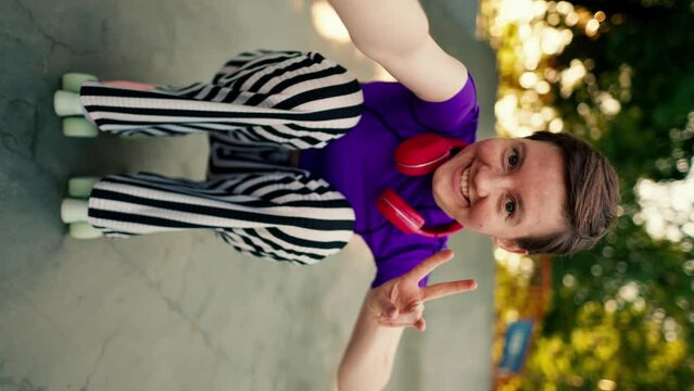 Vertical video: Happy girl with a short haircut in a purple top and red headphones in striped pants rides sitting on pink roller skates in a skate park in summer