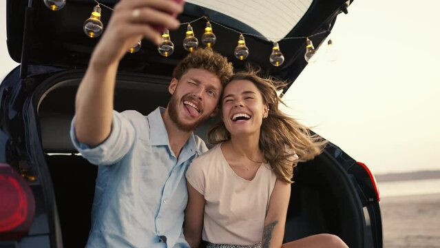 A bearded guy with curly hair in a blue shirt is smiling, grimacing at the camera with his blonde girlfriend in a white top and takes a funny selfie in the trunk of a black car decorated with light