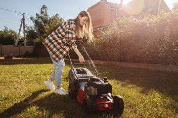 Female gardener working in autumn, cutting grass in backyard. Concept of gardening, work, nature. Housework, gardening and country life. Home garden grass cutting woman mowing with lawn mower. - obrazy, fototapety, plakaty