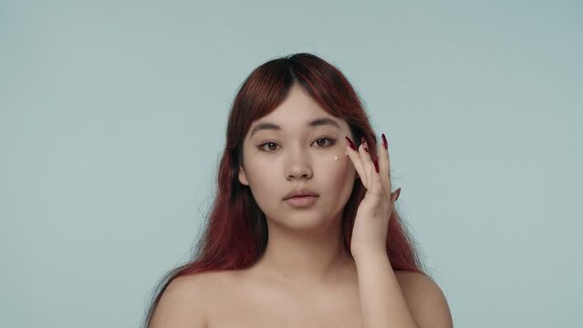 Close-up isolated slow motion video of a young seminude woman with red dyed hair and nude makeup looking at the camera, applying three dots of cream on her cheeck.