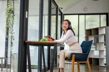 Young Asian woman wearing headphones sitting with legs raised on a chair enjoying using a tablet, watching movies, listening to music in a public work area.